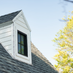 Witte dakkapel met raam op schuin dak van een woning, tegen een blauwe lucht met bomen op de achtergrond.